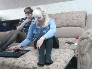 Video of two women on a beige sofa, one wearing a light blue shirt and black tights, typing on a keyboard, the other in glasses, dark hair, and a plaid shirt, reading a book.