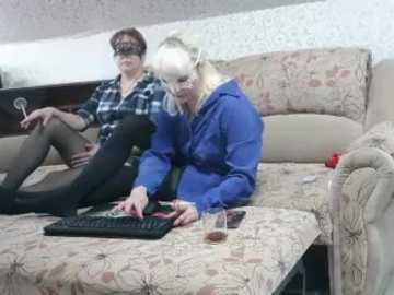 Video of two women on a beige couch, one in a black-and-white plaid shirt, the other in a blue hoodie, playing a video game on a TV.