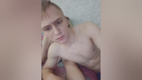 A video of a young, lean, fair-skinned man with short, light brown hair, lying shirtless on a bed with a patterned, multicolored blanket. His face displays a neutral expression. The background features a textured, light-colored wall.
