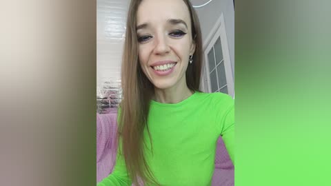 Video of a young Caucasian woman with long, straight brown hair, wearing a bright green long-sleeve shirt, smiling in a well-lit, modern room with white walls, a brick wall, and a pink sofa.