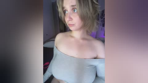 A video of a young woman with short, messy blonde hair, wearing a grey off-shoulder crop top that reveals her bare breasts, in a dimly lit room with purple lighting and a bed visible in the background.