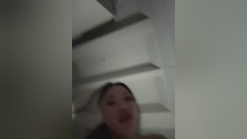 A blurred video shows a woman with dark hair, possibly Asian, wearing a red lipstick, standing in a dimly lit room with white ceiling panels and a minimalistic design.