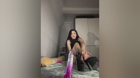 Video of an Asian woman with long black hair, wearing a black crop top and black stockings, sitting on a bed with a purple vibrator in her hand, in a dimly lit, sparsely furnished bedroom.
