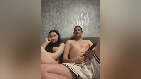 Video of an East Asian couple, nude, sitting on a grey couch against a textured grey wall, man with a beer bottle, woman resting head on hand, casual, intimate setting.