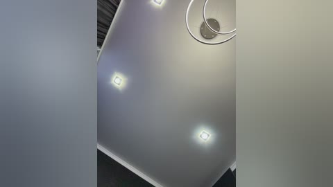 Video of a modern, dimly lit room with a glossy white ceiling featuring circular recessed lights, a silver lamp hanging from the ceiling, and a dark, textured wall in the background.