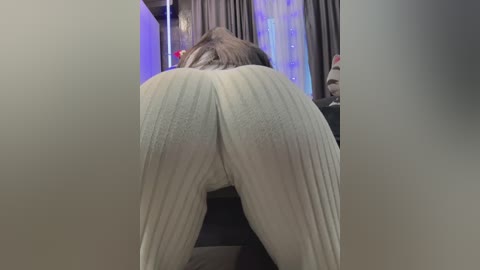 Video of a person with light skin, wearing white ribbed leggings, bent over, viewed from behind, in a dimly lit room with curtains and blue lights.