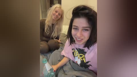 Video of two women, one with long blonde hair and the other with dark hair, both smiling, sitting on a bed. The blonde woman wears black clothing, and the dark-haired woman has a pink graphic tee.