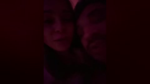 A dimly-lit video showing two young women with light skin and dark hair, their faces close together, creating a sense of intimacy and connection. The background is dark and blurry, adding to the moody atmosphere.