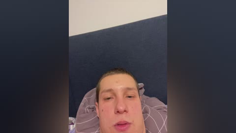 Video of a young Caucasian man with light skin, short hair, and a neutral expression lying on a dark blue and light blue checkered blanket.