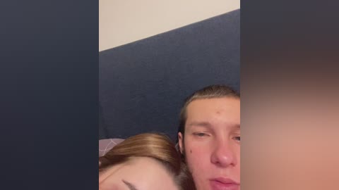 Video of a man with short brown hair sleeping, partially covered by a woman with long blonde hair, in a bed with dark blue and light beige walls.