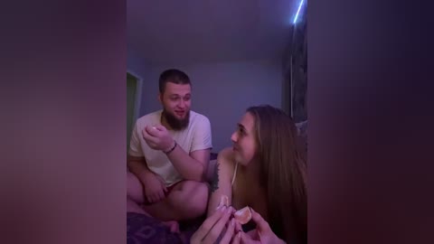 A dimly lit video shows a bearded man in a white t-shirt, sitting on a bed, gesturing while a topless woman with long hair looks at him.