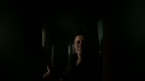 A dimly lit, moody video of a man in a dark suit, holding a cigarette, standing in a narrow, dimly lit hallway. The background is dark and shadowy, emphasizing a tense, possibly sinister atmosphere.