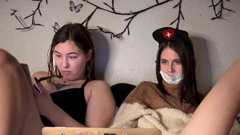 Video of two young women, one with a butterfly tattoo, lying on a bed; one wearing a nurse hat, the other gagged with a cloth.
