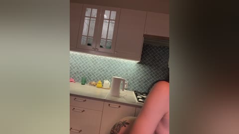 A video captures a modern kitchen with light cabinets, a white countertop, a blue diamond-patterned backsplash, and a person with a tan, muscular build partially visible.