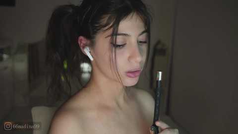 Video of a young woman with light skin and dark hair in a ponytail, listening to headphones, wearing a strapless top. Background is blurry, indoor setting.