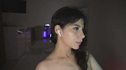 Video of a young woman with dark hair, wearing white earbuds, in a dimly lit room with a faint purple glow and a visible bathroom sink in the background.