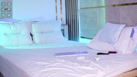 Video of a minimalist, modern bedroom with a white bed, large pillows, and a laptop on the bedspread. The background features a partially visible window with vertical blinds and a wall with a geometric pattern.