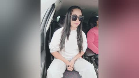 Video of a woman with long black hair, wearing sunglasses, white top, and loose white pants, seated in a car with another person visible in the background.