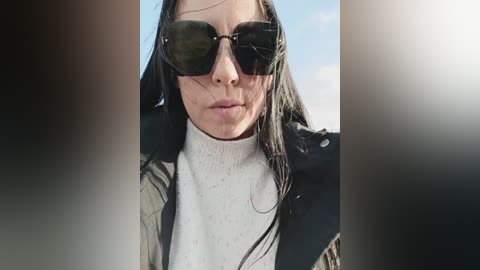Video of a woman with long, wet hair, wearing black sunglasses and a white turtleneck under a black jacket, against a bright blue sky.