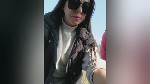 Video of a woman with long black hair, wearing black sunglasses, a grey turtleneck, and a black jacket, outdoors with a clear blue sky in the background.