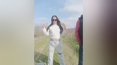 Video of a woman with long dark hair, wearing a white sweater, high-waisted light-wash jeans, and sunglasses, posing on a grassy path under a clear blue sky.