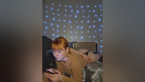 A video of a young woman with straight, reddish-brown hair, wearing a brown sweater, lying on a bed with a gray blanket, focused on her smartphone, surrounded by a string of blue fairy lights in the background.