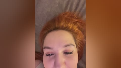 Video of a fair-skinned woman with long, vibrant red hair, lying on a bed. She has closed eyes, defined eyebrows, and subtle makeup. The background features a blurred beige pillow.