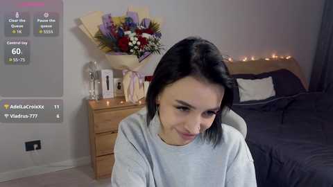 Video of a young woman with short black hair, wearing a grey sweater, sitting in a cozy bedroom with a wooden dresser, floral arrangement, and a neatly made bed.