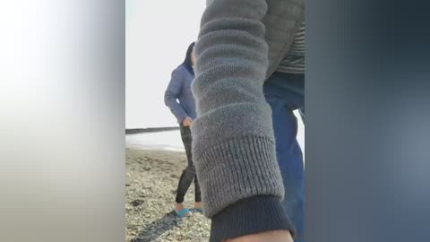 A video showing a person in a grey sweater, walking on a gravelly ground, with a blurred figure in blue clothing in the background.