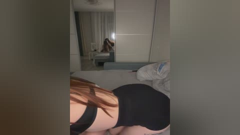 A video of a woman with long red hair, wearing black lingerie, bending over a bed. A mirror reflects her in a dimly lit, modern bedroom with white walls and a closet.