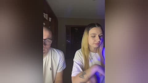 Video of a young woman with long blonde hair and a tattoo on her arm, wearing a white t-shirt, looking at her reflection in a dark mirror.