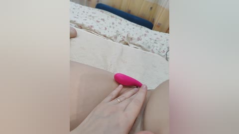Video of a nude woman lying on a bed with a pink vibrator on her vulva. Her fair skin contrasts with the beige sheets.