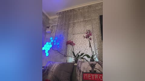 Video of a cozy living room with a gray sofa, a white table with red \"METRO\" sign, blue hexagonal LED lights, and a flower vase with pink orchids on a textured wall.