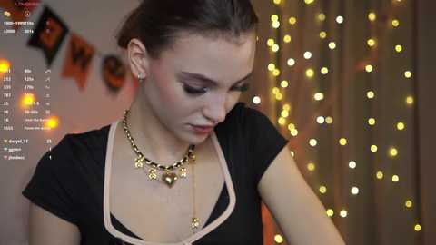 Video of a young Caucasian woman with fair skin and brown hair in a bun, wearing a black top with a gold necklace, in a dimly lit room with string lights and a festive background.