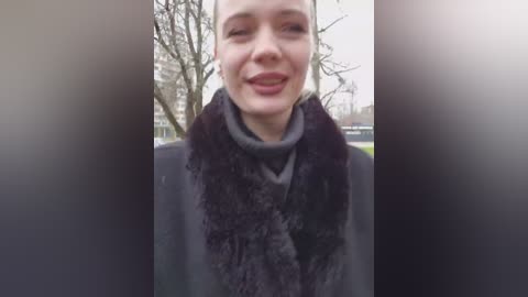 A video shows a smiling woman in a dark, furry coat, standing outdoors in a city park. The background features bare trees and distant buildings.