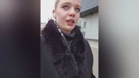 Video of a fair-skinned woman with short blonde hair, wearing a black fur-trimmed coat, looking surprised, standing indoors next to a white wall and black counter.