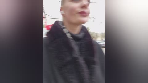 A blurry, low-quality video of a woman in a black coat with a patterned scarf, standing outdoors against a blurred background.
