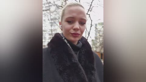 Video of a fair-skinned, blonde woman with a serious expression, wearing a dark coat and fur-trimmed scarf, standing in front of bare trees and a blurred building.