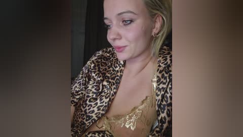 Video of a fair-skinned, blonde woman with shoulder-length hair, wearing a leopard-print jacket and a gold lace bra, sitting against a dark background. She has a subtle smile.