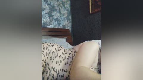 A video of a person lying on a bed with a leopard-print duvet, visible from the waist up. The background features a dark floral wallpaper and a wooden headboard.
