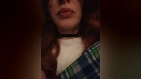 A video of a woman with fair skin and long auburn hair, wearing a black choker and a white ribbed top under a green and blue plaid shirt, set against a dark, blurred background.
