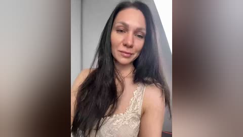 Video of a light-skinned woman with long, straight black hair, wearing a beige lace tank top, standing indoors against a blurred background.