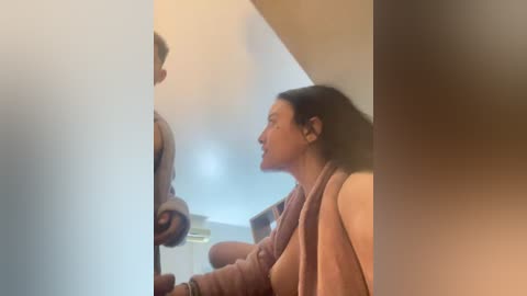 Video of a woman with long dark hair, wearing a beige sweater, standing beside a man wearing a jacket, in a foggy, dimly lit room.