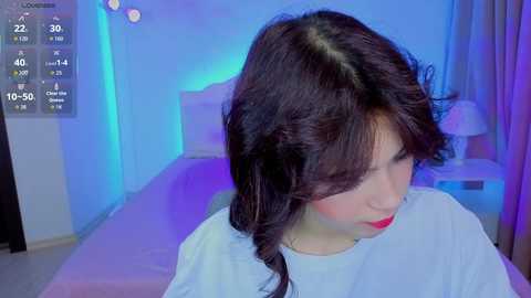 A video of a young woman with dark hair, wearing a white shirt, sitting on a bed in a dimly lit room with blue lighting.