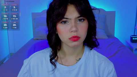 Video of a young woman with dark hair, wearing a white T-shirt, standing in a dimly lit bedroom with blue lighting.