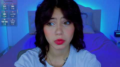 Video of a young woman with wavy dark hair, wearing a white shirt, sitting on a bed with blue lighting, looking surprised.