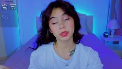 A video of a young woman with long dark hair, fair skin, and a small tattoo, wearing a white shirt, lying on a bed with a purple sheet. The background features a blue-lit room with a bedside table.