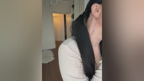 Video of a woman with long black hair tied back, wearing a white long-sleeve top and dangling earrings, standing in a modern, brightly lit living room with hardwood floors and white walls.