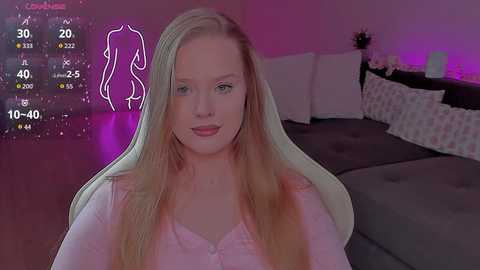 A video of a blonde woman in a white top, sitting in a pink chair with a purple glow, in a cozy bedroom with a grey bed and white pillows.