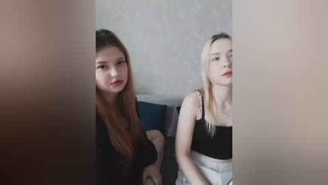 Video of two young women seated on a couch, one with long red hair, the other with long blonde hair, wearing casual black tops. Background shows a plain, light-colored wall.